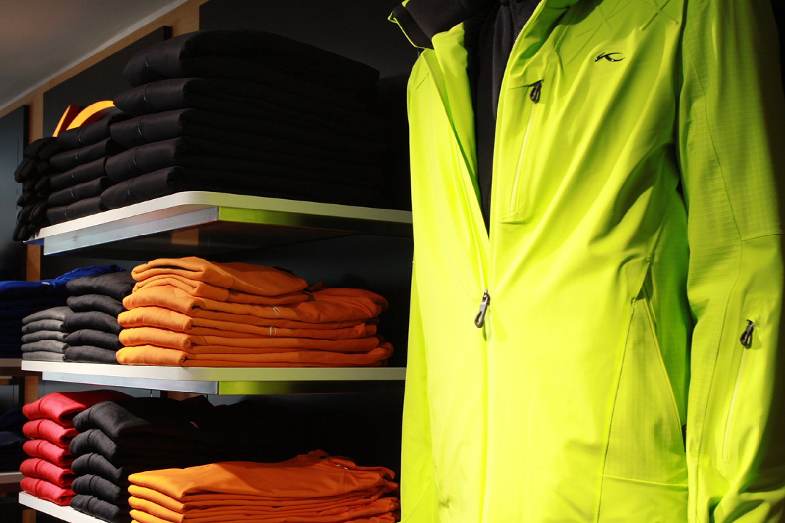 Nahaufnahme auf Regal mit orangen, schwarzen und roten Shirt, so wie einer quietsch grünen Jacke am rechten Rand-alles von der Marke KJUS